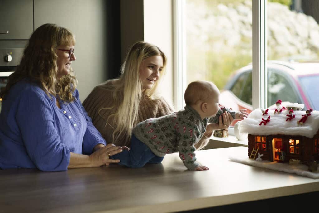 Bilde som viser babyen Mio som strekker seg mot et pepperkakehus. Ved siden av står mamma Tanya og bestemor Marianne.