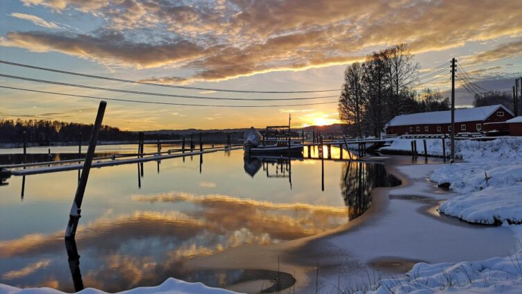 En vakker solnedgang over en snødekt båthavn. Vannet er stille og reflekterer den gyldne himmelen, mens noen båter er fortøyd langs brygga.