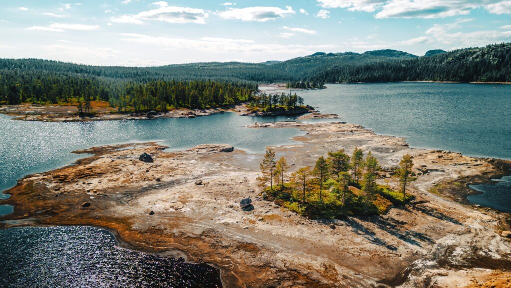Et vakkert kystlandskap med innsjøer og små øyer omgitt av skog. Fjell og skoger i det fjerne under en delvis skyet himmel.