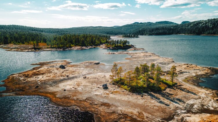 Et vakkert kystlandskap med innsjøer og små øyer omgitt av skog. Fjell og skoger i det fjerne under en delvis skyet himmel.