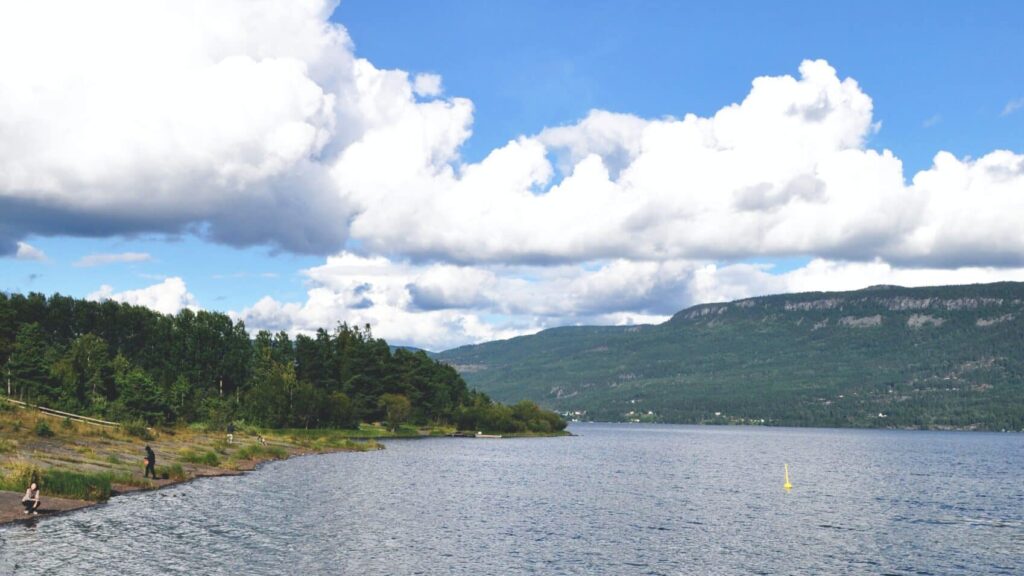 Utsikt over en innsjø omgitt av skogkledde fjell under en klar blå himmel med noen skyer.