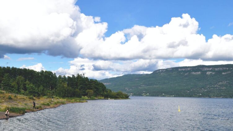 Utsikt over en innsjø omgitt av skogkledde fjell under en klar blå himmel med noen skyer.