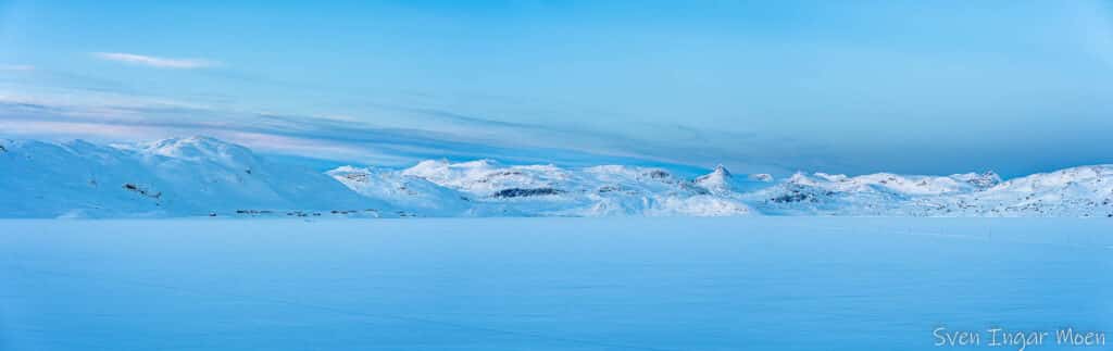 Snødekt fjelllandskap og vann under en klar blå himmel.