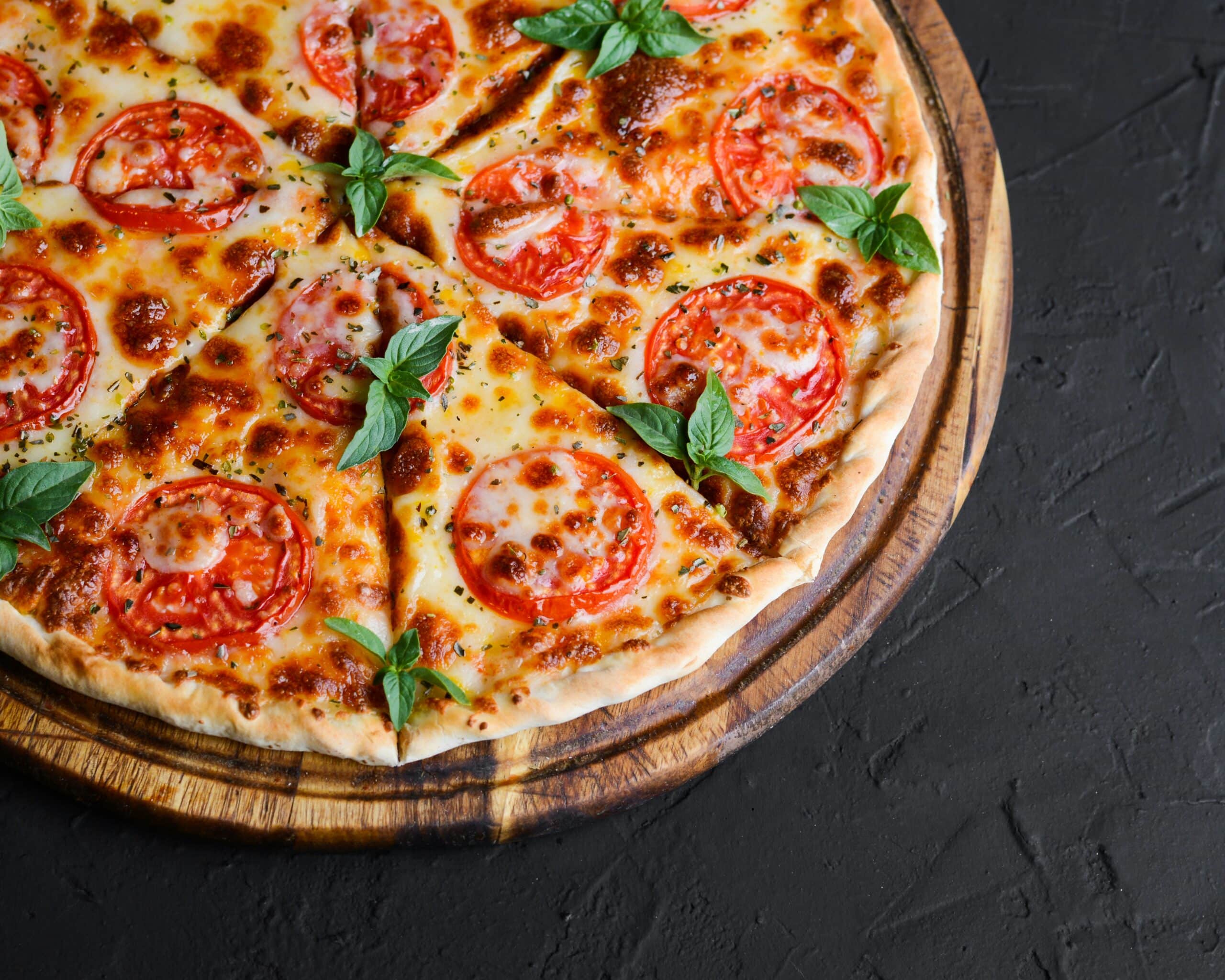 En stekt pizza på et rundt trefat, toppet med tomatskiver, ost og basilikumblader. Pizzaen er delt i stykker, og bildet har en mørk bakgrunn som fremhever maten.