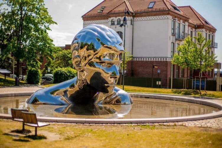 Skinnende metallskulptur i en park, formet som et menneskehode.